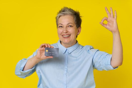 Holding credit, credit card grey haired woman gesturing OK with other hand making online payment or shopping. Pretty mature woman in blue shirt isolated on yellow background. Banking concept.