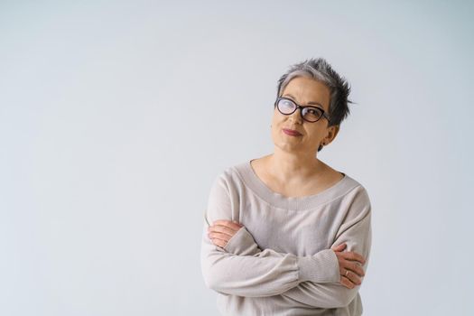 Serious slightly smiling mature grey hair woman in glasses posing with hands folded looking at camera wearing white blouse, copy space isolated on white background. Healthcare, aged beauty concept.