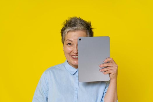 Grey haired mature woman with hide behind tablet pc working online or social media. Pretty woman in 50s wearing blue blouse isolated on yellow. Mature people and technologies.