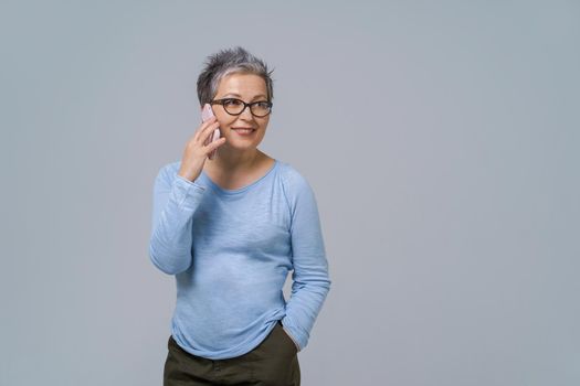 Talking on the phone mature grey hair woman in glasses turn sideways holding smartphone in her hand wearing blue blouse and black skirt. Pretty woman in blue shirt isolated on white background.