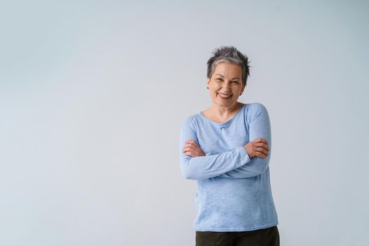 Charming positive emotions mature grey hair woman posing with hands folded looking at camera wearing blue blouse, copy space isolated on white background. Healthcare, aged beauty concept.