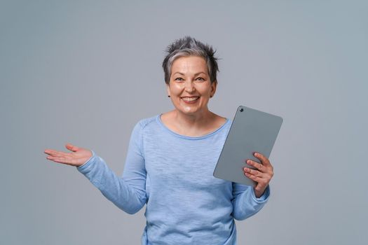 Happy middle age woman with grey hair and tablet pc in hands browsing, working, shopping online. Pretty woman in 50s wearing blue blouse isolated on grey. Mature people and technologies.