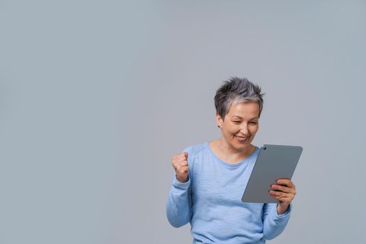 Excited mature woman with grey hair looking at tablet pc shopping, working online or having a video call. Pretty woman in 50s wearing blue blouse isolated on grey. Mature people and technologies.