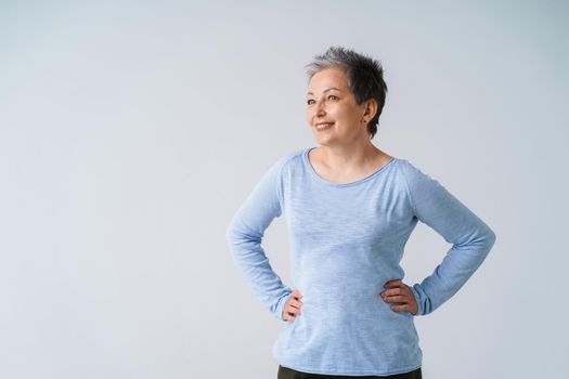 Charming mature silver, grey hair woman posing with hands on her hips looking sideways away wearing blue blouse, isolated on white background. Healthcare, aged beauty concept. Copy space.