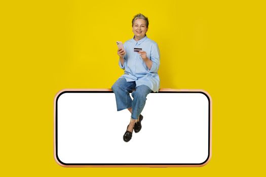Silver, grey haired woman with phone and debit, credit card in hand making online payment or shopping sitting on a huge, giant smartphone, wearing blue shirt and jeans isolated on yellow background.