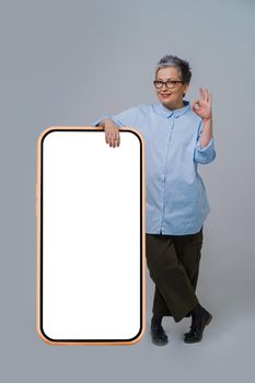 Gesturing OK grey hair mature business woman in glasses leaned on giant, huge smartphone with white screen wearing oversized blue shirt isolated on white background. Free space mock up.