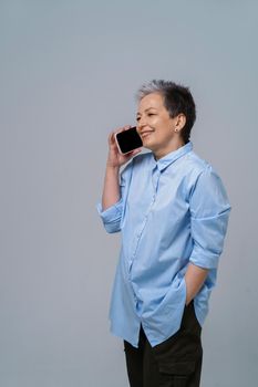 Mature grey hair woman talking on the phone turn sideways holding smartphone in her hand wearing blue shirt and black skirt. Pretty woman in blue shirt isolated on white background.