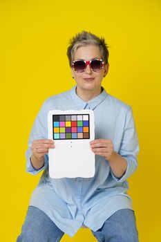 Sitting on a chair mature grey hair woman in UK flag sunglasses with color checker in hands looking at camera wearing blue shirt isolated on yellow background. Healthcare, aged beauty concept.