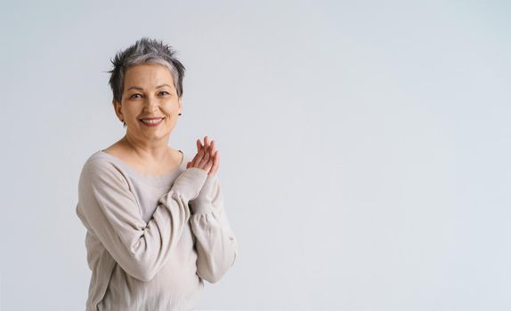 Mature grey hair woman posing with hands folded smiling looking at camera wearing white blouse, copy space on right isolated on white background. Healthcare concept. Aged beauty concept.