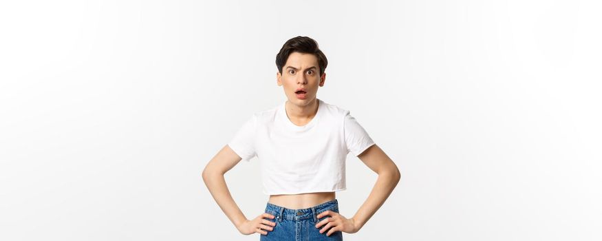 Shocked and confused gay man staring at camera startled, wearing crop top, holding hands on waist, standing over white background.