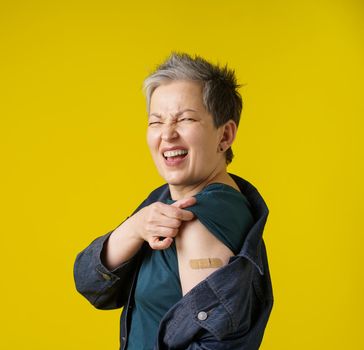 Grey hair senior woman showing shoulder with band aid after having shot of vaccine wearing trendy clothes isolated on yellow background. Mature woman 50s vaccination and healthcare concept.