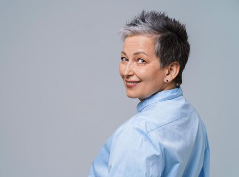 Grey hair mature woman posing standing back half turn looking over shoulder at camera and smile wearing blue shirt isolated on white background. Aged beauty. Copy space.