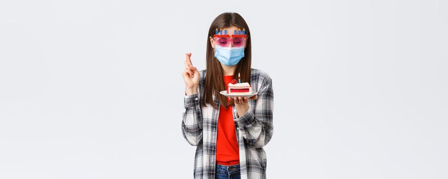 Coronavirus outbreak, lifestyle social distancing and celebration concept. Enthusiastic happy girl in glasses and medical mask celebrate birthday on quarantine, cross fingers, make wish at cake.