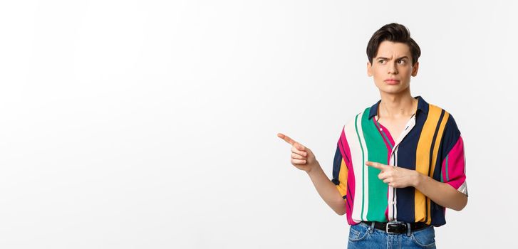 Suspicious young man looking and pointing fingers left with doubtful face, standing unsure in summer shirt, white background.