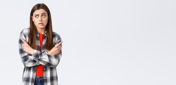 Lifestyle, different emotions, leisure activities concept. Indecisive silly cute girl facing hard choice, cross hands and pointing sideways, leaning to left variant, biting lip confused and puzzled.