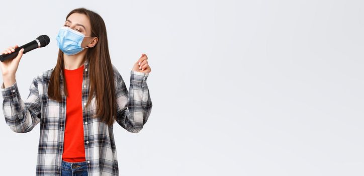 Coronavirus outbreak, leisure on quarantine, social distancing and emotions concept. Carefree young girl in medical mask, singing with microphone, close eyes, enjoying karaoke.