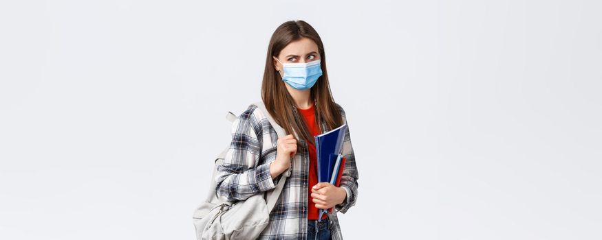 Coronavirus pandemic, covid-19 education, and back to school concept. Displeased female student in medical mask and backpack, look behind at upper left corner with disdain or dismay, hold notebooks.