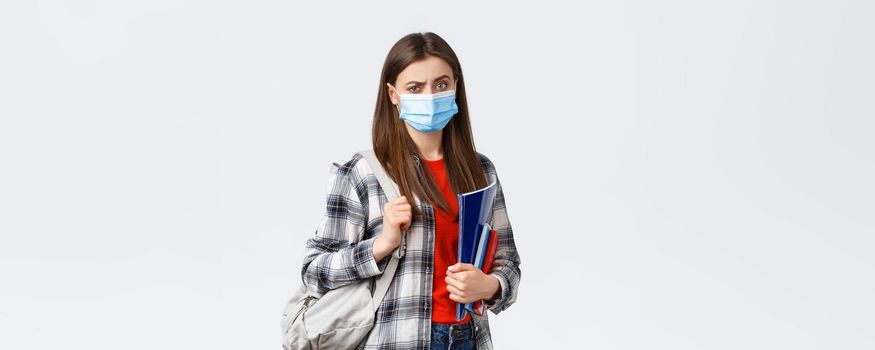 Coronavirus pandemic, covid-19 education, and back to school concept. Doubtful and skeptical female student in medical mask frowning disappointed, hold notebooks and backpack.