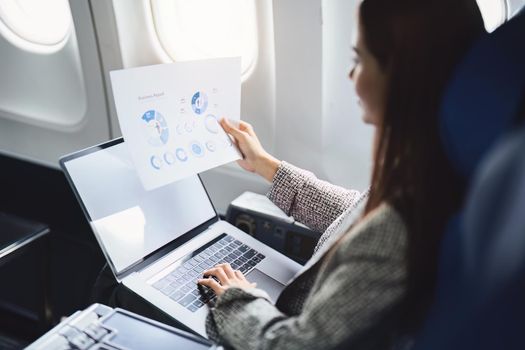 A successful asian business woman or female entrepreneur in formal suit in a plane sits in a business class seat and uses a documents with computer laptop during flight.