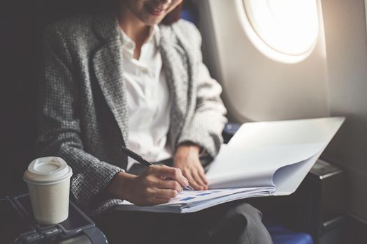 A successful asian business woman or female entrepreneur in formal suit in a plane sits in a business class seat and uses a pen with documents for work during flight.