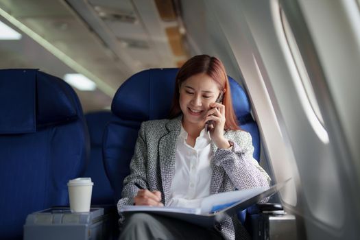 portrait of A successful asian business woman or female entrepreneur in formal suit in a plane sits in a business class seat and uses smartphone with documents for work during flight.