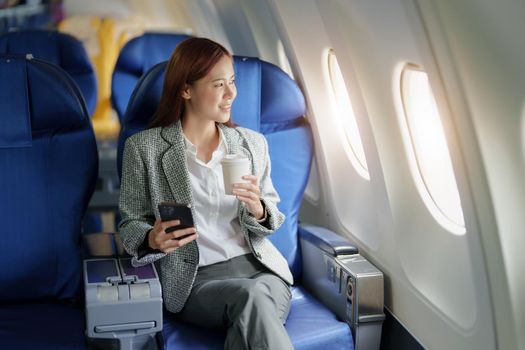 portrait of A successful asian business woman or female entrepreneur in formal suit in a plane sits in a business class seat and uses a smartphone with drink coffee during flight, relax concept.