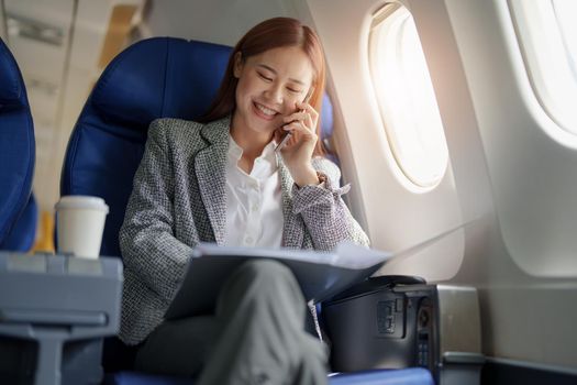 portrait of A successful asian business woman or female entrepreneur in formal suit in a plane sits in a business class seat and uses smartphone with documents for work during flight.