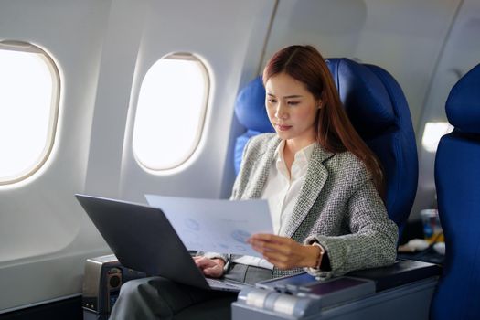 portrait of A successful asian business woman or female entrepreneur in formal suit in a plane sits in a business class seat and uses a documents with computer laptop during flight.
