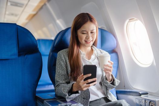 portrait of A successful asian business woman or female entrepreneur in formal suit in a plane sits in a business class seat and uses a smartphone with drink coffee during flight, relax concept.