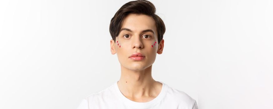 People, lgbtq and beauty concept. Close-up of beautiful androgynous man with glitter on face, looking at camera, standing over white background.