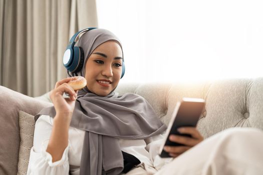 Beautiful Asian muslim woman using mobile phone while sitting on sofa, girl doing chat text message on smartphone and smiling.