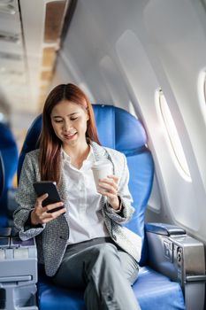 portrait of A successful asian business woman or female entrepreneur in formal suit in a plane sits in a business class seat and uses a smartphone with drink coffee during flight, relax concept.