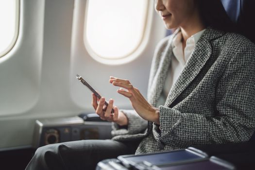 A successful asian businesswoman or female entrepreneur in formal suit in a plane sits in a business class's seat and uses a smartphone during flight.
