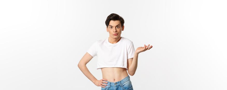 Handsome androgynous man in crop top looking confused, shrugging clueless, standing over white background.
