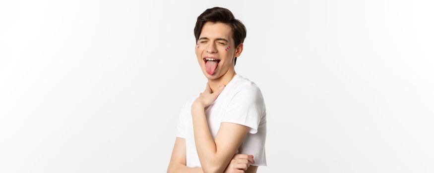 People, lgbtq and beauty concept. Close-up of carefree gay man with glitter on face, showing tongue and making funny face, standing over white background.