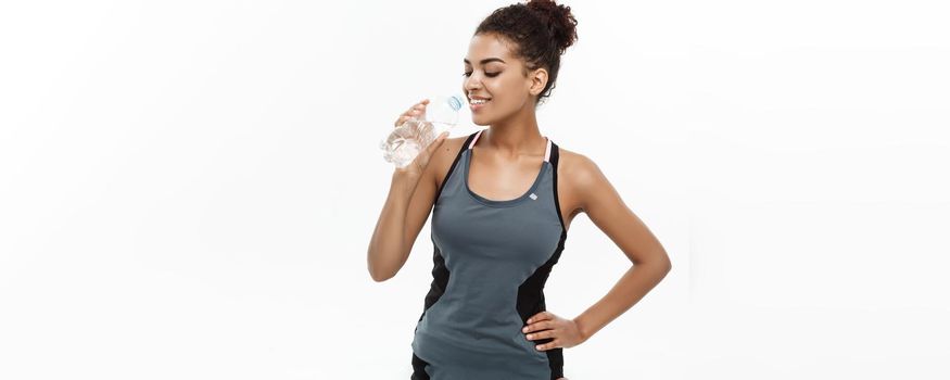 Healthy and Fitness concept - beautiful African American girl in sport clothes drinking water by plastic bottle after workout. Isolated on white studio background.