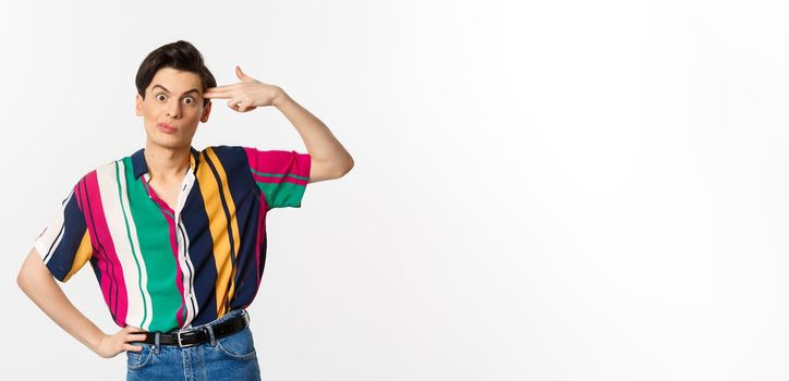 Annoyed gay man making finger gun sign, shooting hismelf from irritation and boredom, standing over white background.