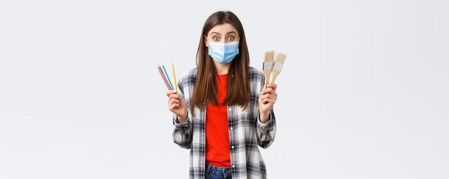 Social distancing, leisure and hobbies on covid-19 outbreak, coronavirus concept. Excited cheerful girlfriend suggest drawing together, holding painting brushes and colored pencils, wear medical mask.