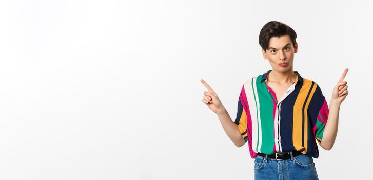 Handsome gay man pointing fingers sideways, showing two variants, demonstrate promos, standing over white background.