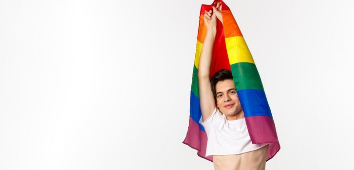 Vertical view of beautiful androgynous gay man raising rainbow flag and smiling happy, standing in crop top and jeans against white background.