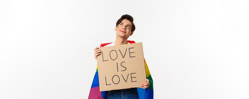 Dreamy young queer person smiling and looking at upper left corner, holding love is love sign for pride parade, wearing Rainbow flag, white background.