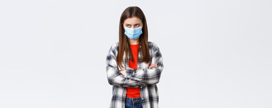 Coronavirus outbreak, leisure quarantine, social distancing and emotions concept. Mad young cute girl in medical mask, cross arms chest, look from under forehead upper left corner, jealous or angry.