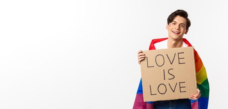 Smiling gay man activist holding sign love is love for lgbt pride parade, wearing Rainbow flag, standing over white background.