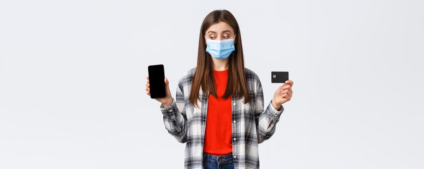 Coronavirus outbreak, working from home, online shopping and contactless payment concept. Girl in medical mask showing mobile phone and credit card, peek at screen making order.