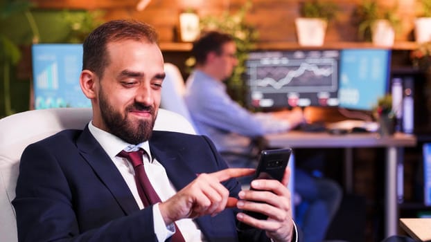 Businessman smiling and looking at his smartphone. Colleague working in the back