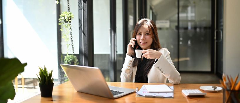 Confident asian businesswoman drinking coffee and talking on mobile phone at office desk.