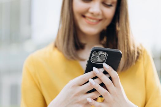 Joyful young attractive european business girl using mobile phone, smiling when reading sms, spending time outdoors. Urban people portraits.