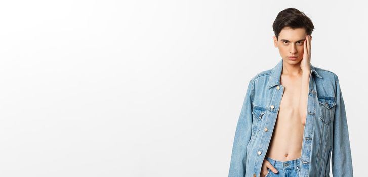 Handsome and sassy gay man wearing denim jacket on bare torso, touching his face and looking confident at camera, standing over white background.