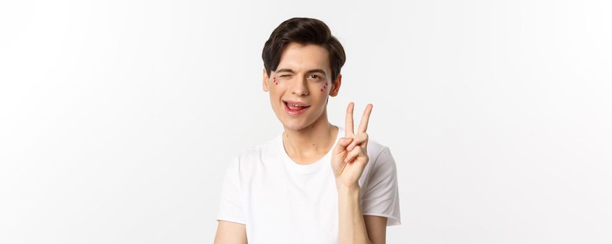 People, lgbtq community and lifestyle concept. Happy and cute gay man with glitter on face, showing peace sign and smiling, celebrating pride holiday, white background.