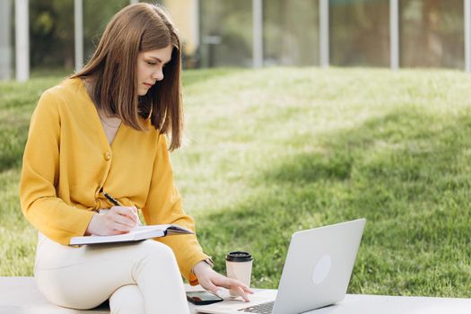 Portrait of focused student girl doing homework next to laptop computer. Teenage girl writing in notebook in front of laptop outdoors. Pretty girl studying at home education.